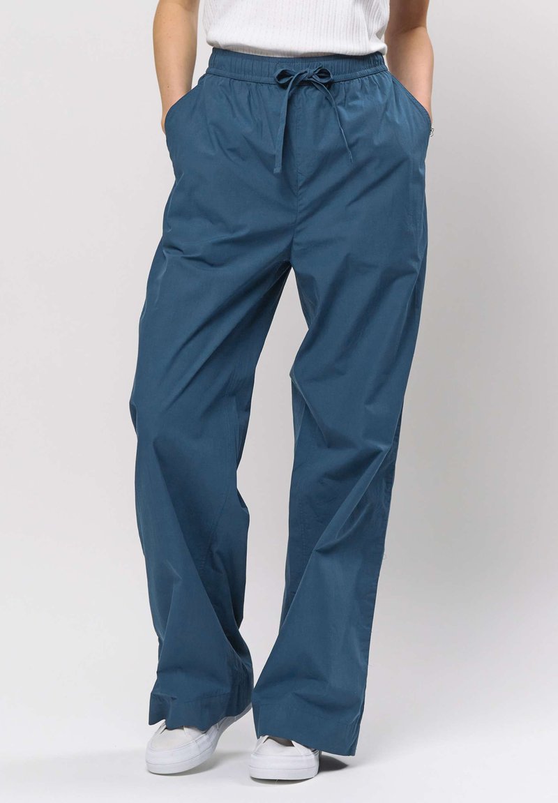 Woman wearing loose blue drawstring pants and white sneakers, hands in pockets, standing against plain light background.