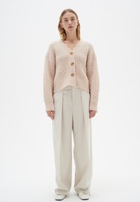 Helles rosa gestricktes Cardigan mit einem cropped Design, V-Ausschnitt, strukturiertem Muster und vier großen Knöpfen. Kombiniert mit beigen, weiten Hosen.