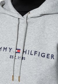 Sudadera gris con cordones, que presenta un logo bordado en azul marino y detalles en rojo y blanco. Textura suave y diseño casual.