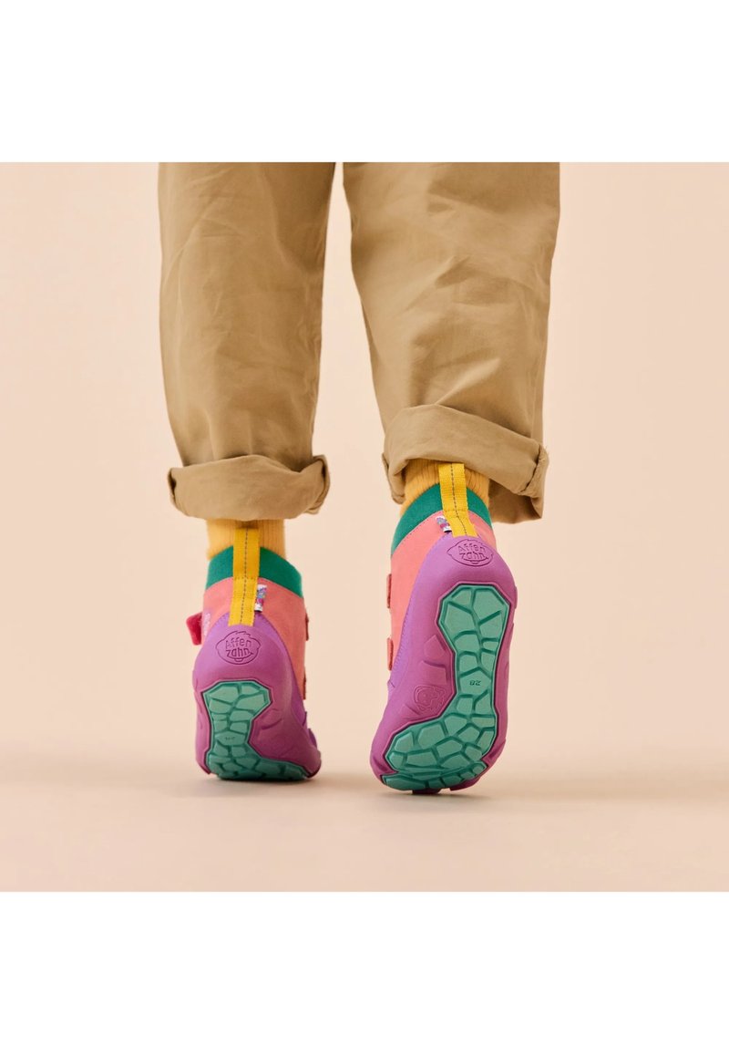 Pieds portant des baskets colorées à semelles violettes et des chaussettes jaunes, debout sur un sol beige avec un pantalon marron retroussé.