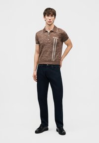 Jeune homme portant un polo en maille à manches courtes marron avec des rayures verticales et un jean noir, debout avec une main derrière le dos.