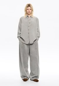 Light gray denim shirt with a button-up front and chest pockets, paired with loose gray trousers. Brown suede slip-on shoes complete the look.