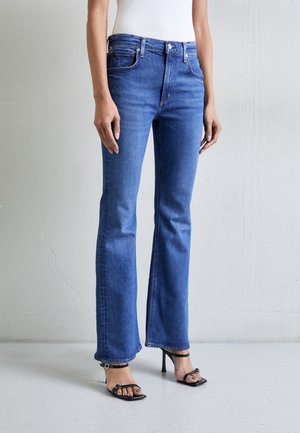 Femme portant un jean bootcut bleu et des sandales noires à talons hauts, debout contre un mur uni gris clair.