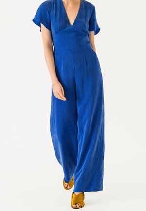 Niebieski lniany kombinezon z szerokimi nogawkami, dekoltem w kształcie litery V i krótkimi rękawami. Posiada dopasowaną talię i gładką fakturę, zestawiony z żółtymi sandałami na obcasie.