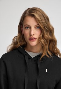 Felpa nera con uno strato interno grigio, cappuccio con coulisse e un piccolo logo bianco sul petto. Tessuto morbido con una superficie liscia.