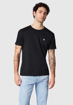 Jonge man met krullend haar, gekleed in een zwart t-shirt en lichtblauwe spijkerbroek, toont tatoeages op beide onderarmen tegen een effen achtergrond.