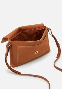 Brun mockaskinn crossbody-väska med öppet lock, framficka med guldknapp och justerbar läderrem på vit bakgrund.