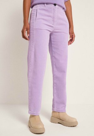 Personne portant un pantalon en velours côtelé violet clair et des bottines beiges, debout devant un fond clair uni.