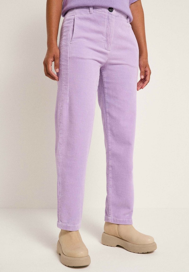 Personne portant un pantalon en velours côtelé violet clair et des bottines beiges, debout devant un fond clair uni.