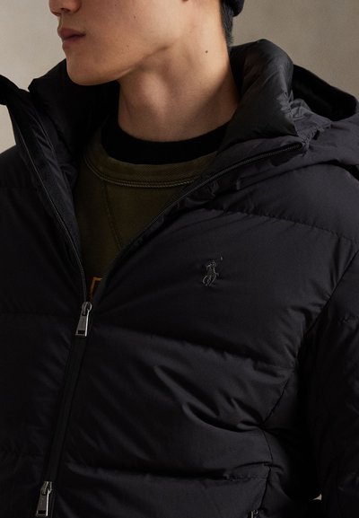 Črna puhovka z visokim ovratnikom, z zadrgo in majhnim vezenim logotipom na prsih. Topel material s prepihanom teksturo.