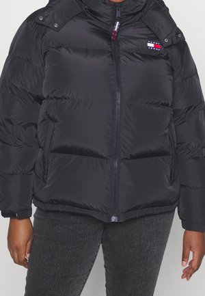 Sort puffert jakke med lynlås foran og Tommy Jeans-logo på brystet, båret med mørkegrå bukser på en person med brun hud.