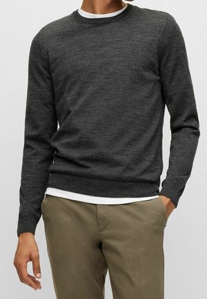 Mand iført mørkegrå crewneck trøje over hvid skjorte, parret med lysebrune bukser, står med den ene hånd i lommen.