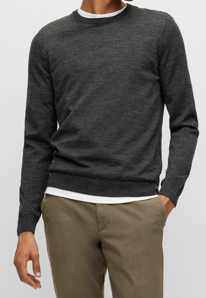 Homme portant un pull gris foncé à col rond par-dessus une chemise blanche, associé à un pantalon marron clair, debout avec une main dans la poche.