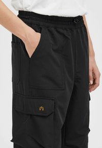Pantaloni cargo neri con vita elastica, tasche laterali e un sottile logo arancione. Il tessuto sembra morbido e leggero.