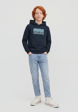 Un garçon roux se tient debout, les mains dans les poches, portant un sweat à capuche bleu marine, un jean bleu clair et des baskets montantes blanches sur un fond gris clair.