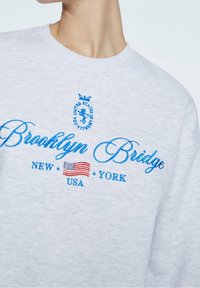Grijze sweatshirt met blauwe geborduurde tekst "Brooklyn Bridge," "New York," en "USA," met een klein ontwerp van de Amerikaanse vlag.