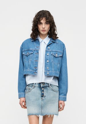 Giovane donna con capelli ricci che indossa una camicia azzurra con bottoni, una giacca di jeans blu corta e una gonna di jeans sbiadita con orlo sfrangiato.