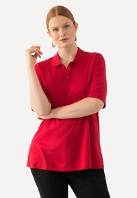 Camisa polo roja de mangas cortas, con tres botones y corte recto. El material parece suave y liso. Pantalones negros que se llevan con ella.