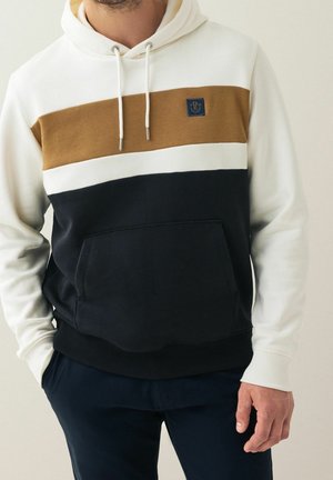 Homme portant un sweat à capuche color-block avec manches blanches, bande marron sur la poitrine, partie inférieure noire et poche avant, debout avec une main dans la poche.