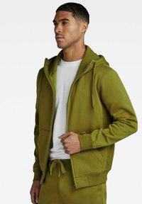 Felpa con zip di colore verde oliva realizzata in tessuto morbido, con cappuccio regolabile, tasca a canguro e polsini a costine. Indossata sopra una maglietta bianca.
