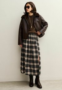 Veste en cuir marron avec col en fourrure, pull marron, jupe à carreaux avec fente latérale, bottes marron jusqu'aux genoux et lunettes de soleil sombres.