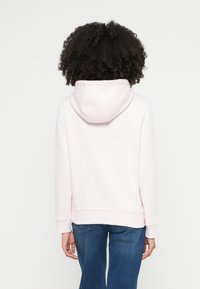 Jasnoróżowa bluza z kapturem z kieszenią kangurka, sznurkiem regulującym kaptur oraz ściągaczami na rękawach, w połączeniu z niebieskimi dżinsami, widziana od tyłu.