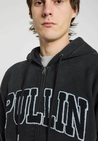 Haut de sweatshirt zippé noir avec "PULL.IN" en lettres blanches et texturées. Le matériau est doux, avec une capuche à cordon de serrage et un zip contrasté.