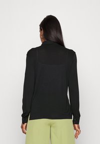 Svart långärmad skjorta med krage, med en slät yta och knappdetaljer på ärmsluten. Bakstycket har en diskret genomskinlig panel.