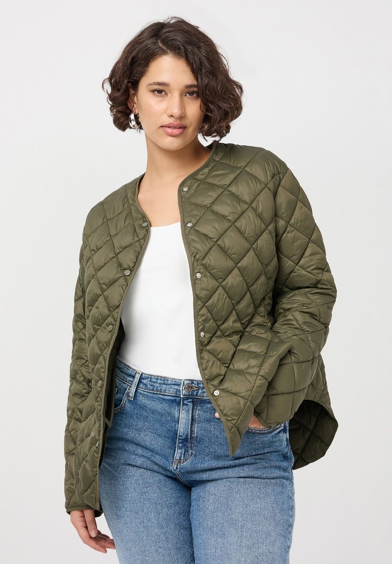 Giacca trapuntata verde oliva con collo arrotondato, bottoni a pressione e fessure laterali, abbinata a una maglietta bianca e jeans di denim.