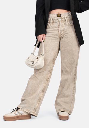 Beige wijde jeans met een verweerd uiterlijk, gecombineerd met een zwart cropped top, een zwarte blazer en lichtgekleurde sneakers, terwijl ze een kleine handtas vasthoudt.