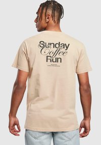 Beige cotton t-shirt with short sleeves, featuring black graphic text on the back: “Sunday Coffee Run” with decorative accents.