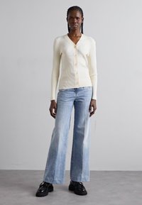 Kremfarget ribbestrikket cardigan med V-hals og knapplukking, kombinert med lyseblå vide jeans og svarte sko. Glatt teksturfinnish.