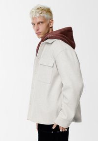 Lichtgrijs wollen jack met een ontspannen pasvorm, voorzien van een knopenrij aan de voorkant, twee borstzakken en een bruine capuchon die eronder zichtbaar is.