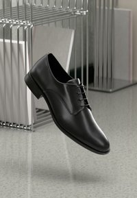 Chaussure habillée en cuir noir avec un bout arrondi, un design à lacets et des coutures minimales, présentée au-dessus d'un sol gris brillant et d'un arrière-plan métallique.