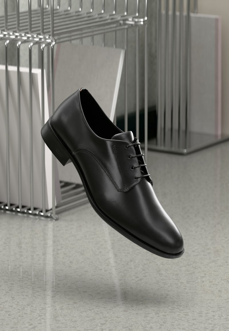 Chaussure habillée en cuir noir avec un bout arrondi, un design à lacets et des coutures minimales, présentée au-dessus d'un sol gris brillant et d'un arrière-plan métallique.
