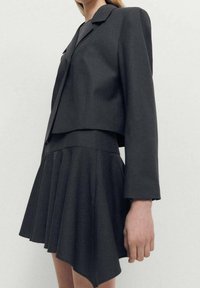 Black tailored blazer paired with a pleated skirt. The fabric is smooth, with a structured design and asymmetrical hemline.