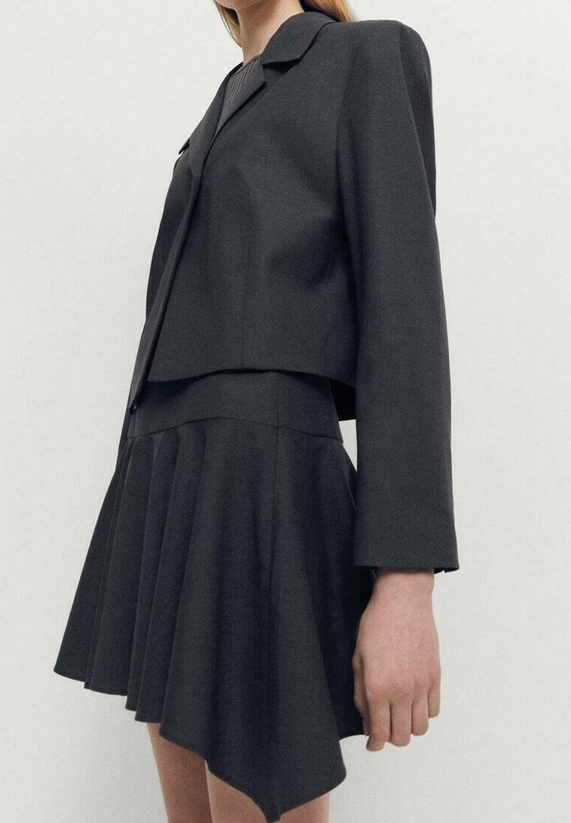 Black tailored blazer paired with a pleated skirt. The fabric is smooth, with a structured design and asymmetrical hemline.