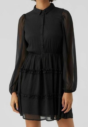 Robe noire à manches longues avec manches bouffantes transparentes, col à boutons, taille élastique et jupe à volants étagée portée par une personne debout.