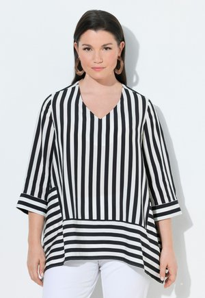 Mujer con una blusa de rayas verticales en blanco y negro, mangas tres cuartos y pantalones blancos, de pie contra un fondo blanco.