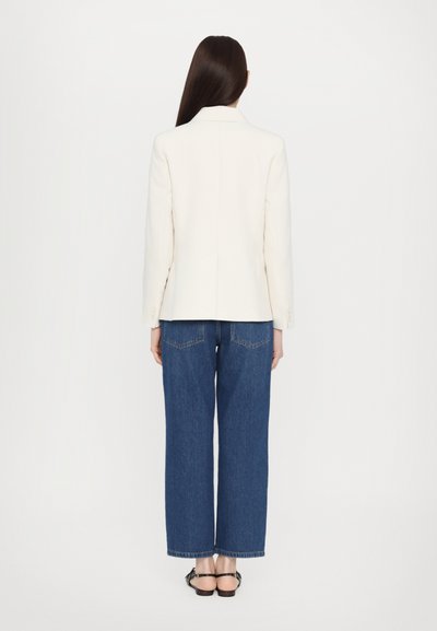 Blazer bianco su misura con spalle strutturate, abbinato a jeans in denim blu a taglio morbido. Il modello indossa sandali neri.