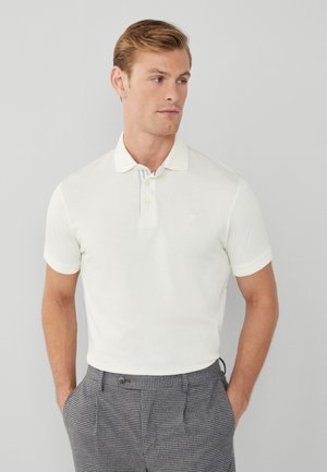 Hvid kortærmet polo-shirt med krave, der har en stribet indre krave og et lille logo på brystet, parret med ternede bukser.