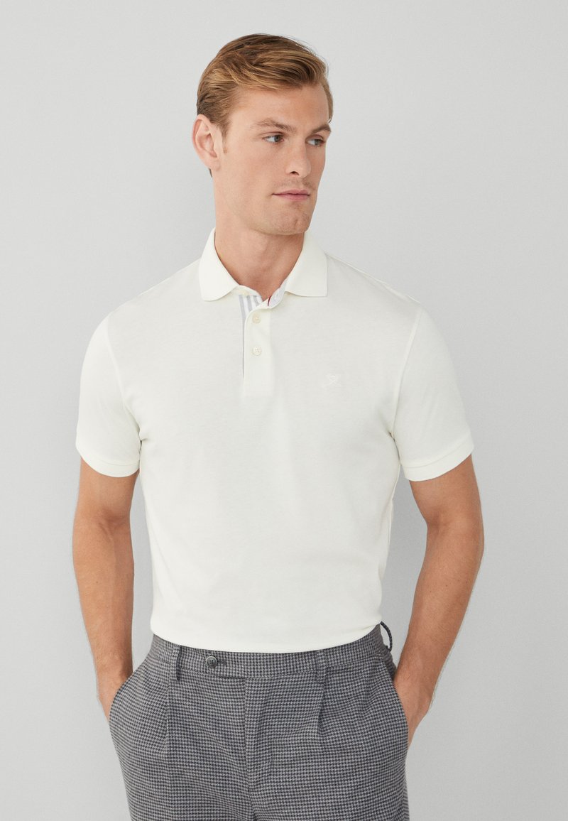 Hvid kortærmet polo-shirt med krave, der har en stribet indre krave og et lille logo på brystet, parret med ternede bukser.