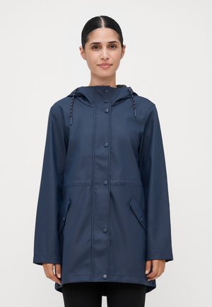 Woman wearing a navy blue long waterproof raincoat with snap buttons and a hood, standing against a white background.