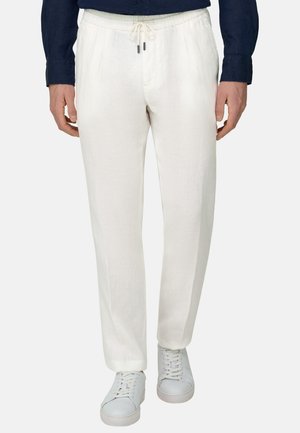 Mężczyzna noszący białe spodnie z troczkiem oraz białe trampki, widoczny od talii do stóp na jednolitym tle.