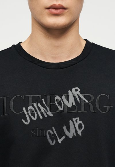 Κοντινή λήψη ενός ατόμου που φοράει μαύρο φούτερ με το κέντημα "ICEBERG" και την έντονη γκρι γραφή "JOIN OUR CLUB" σε στυλ γκράφιτι.