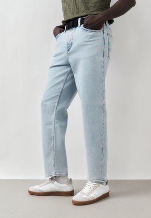 Személy világoskék farmert, fehér gumitalpú tornacipőt és zöld texturált felsőt visel, zsebre tett kézzel áll.