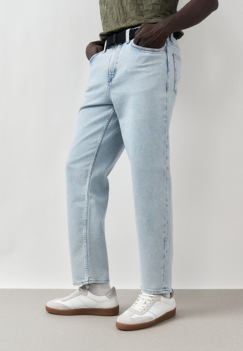 Άτομο που φοράει ανοιχτό μπλε τζιν, λευκά αθλητικά παπούτσια με λαστιχένιες σόλες και πράσινο υφασμάτινο τοπ, στέκεται με τα χέρια στις τσέπες.