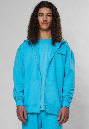 Lys blå zip-up hettegenser laget av myk stoff, med en frontlomme og ribbede mansjetter. Bært over en matchende blå skjorte.
