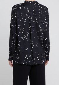 Svart långärmad blus med stjärnmönster. Tillverkad av mjukt tyg, med knäppning vid ärmsluten och en avslappnad passform. Detalj med ok på baksidan ingår.