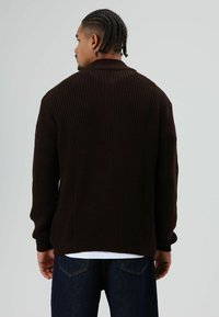 Donkerbruine gebreide trui met een geribbelde textuur, voorzien van een hoge hals en aangesloten manchetten, gedragen over een wit overhemd en een blauwe spijkerbroek.